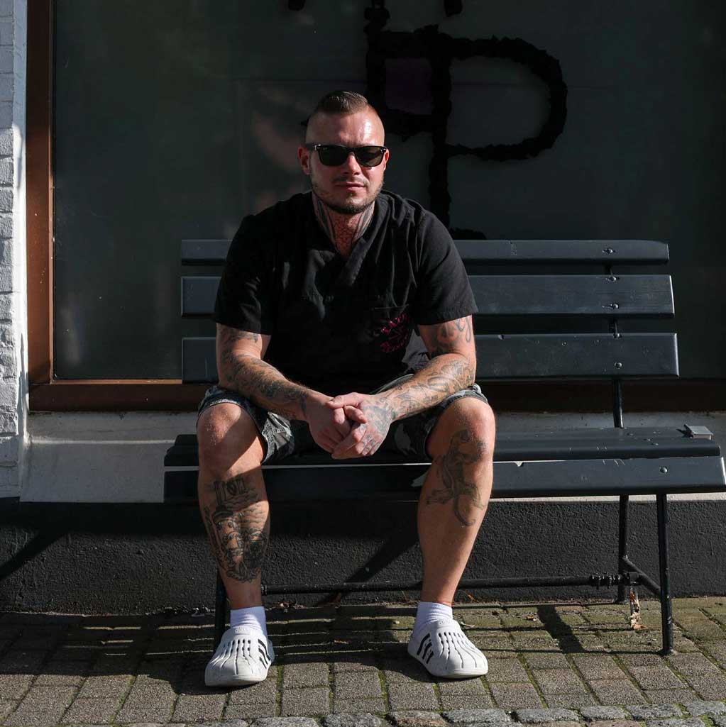 Portrait von Ossi - Tattoo Pleasure, lächelnd mit verschränkten Armen, tätowiertem Arm und kariertem Hemd.